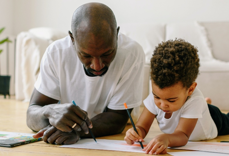 A father and adoptive son lie on the floor together doing colouring