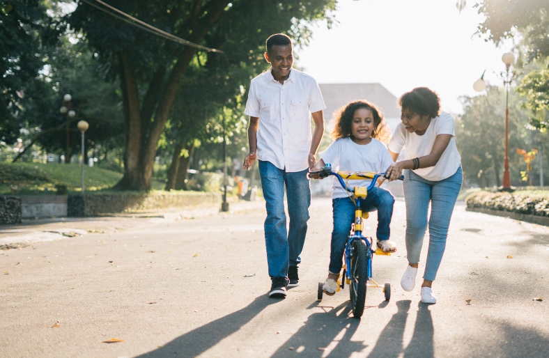 A mum and dad are outside helping to teach their daughter to cycle